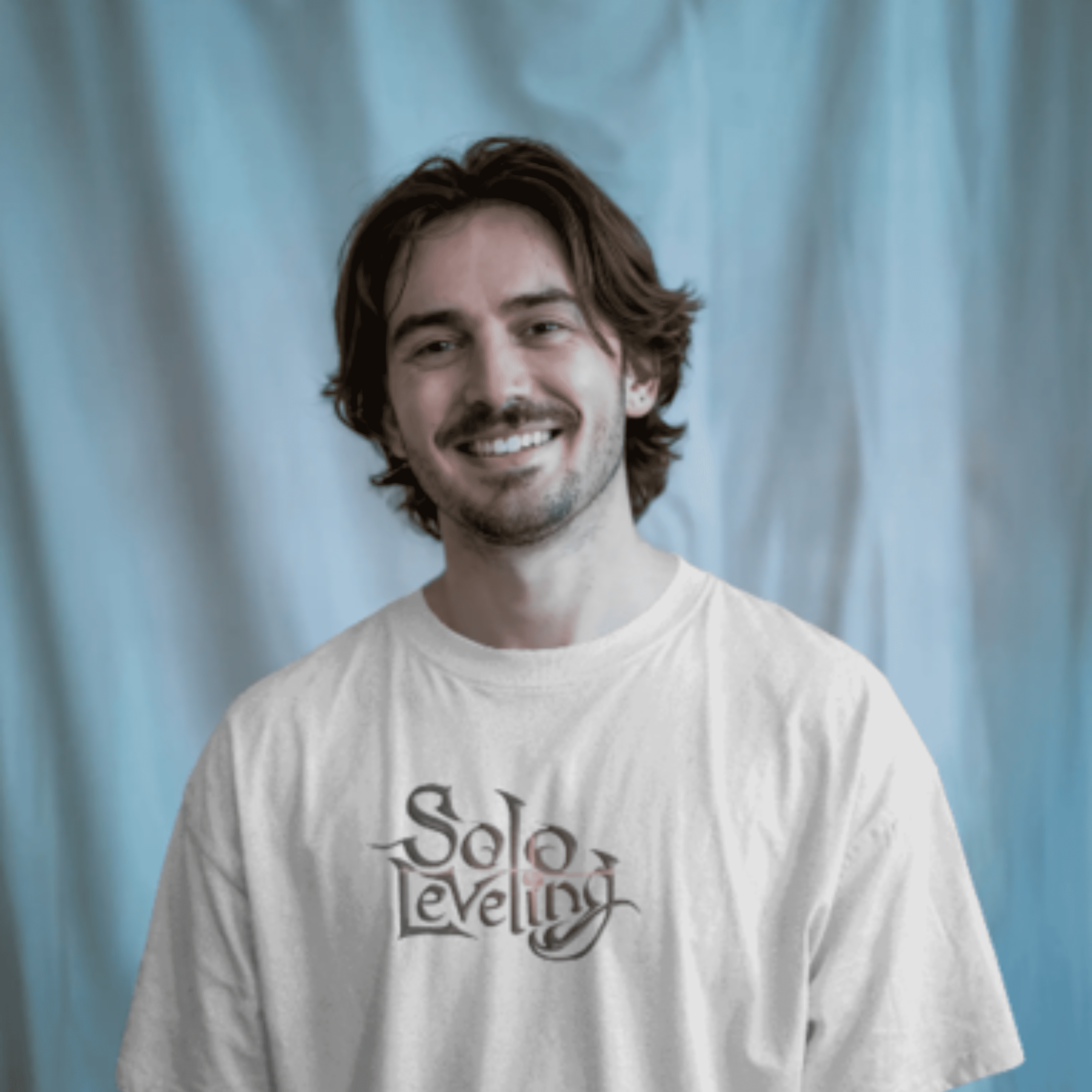 A man wearing a white T-shirt with the text “Solo Leveling” printed on the front in a fantasy-style serif font. He’s smiling confidently against a cool-toned, softly draped background.