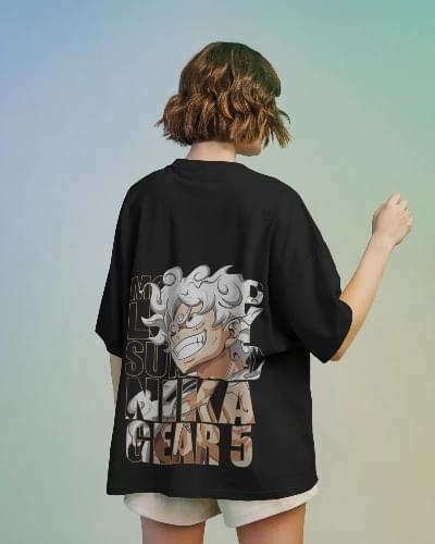 A woman wearing an oversized black T-shirt featuring Luffy in his Gear 5 (Sun God Nika) form from One Piece, with bold text "NIKA GEAR 5" printed on the back. She’s standing against a soft gradient background, holding up a peace sign.