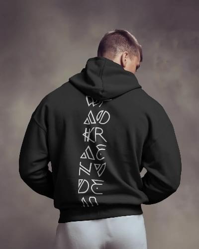 A man wearing a black Wakanda Shadow hoodie with vertically stacked, stylized white geometric letters running down the back. The background is a smoky, gradient grey, giving a moody, mysterious vibe.