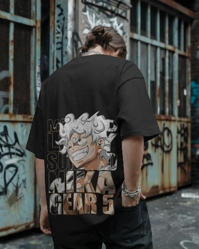 A man wearing a black oversized T-shirt featuring a bold graphic of Luffy in Gear 5 (Nika form) from One Piece, with graffiti-covered urban walls in the background.